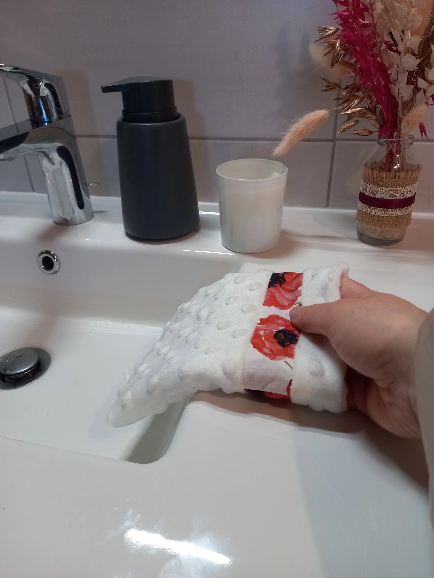 Gant démaquillant lavable en minky blanc à pois en relief et bande à coquelicots rouges, enfilé sur une main inclinée vers la vasque, dans une salle de bain.