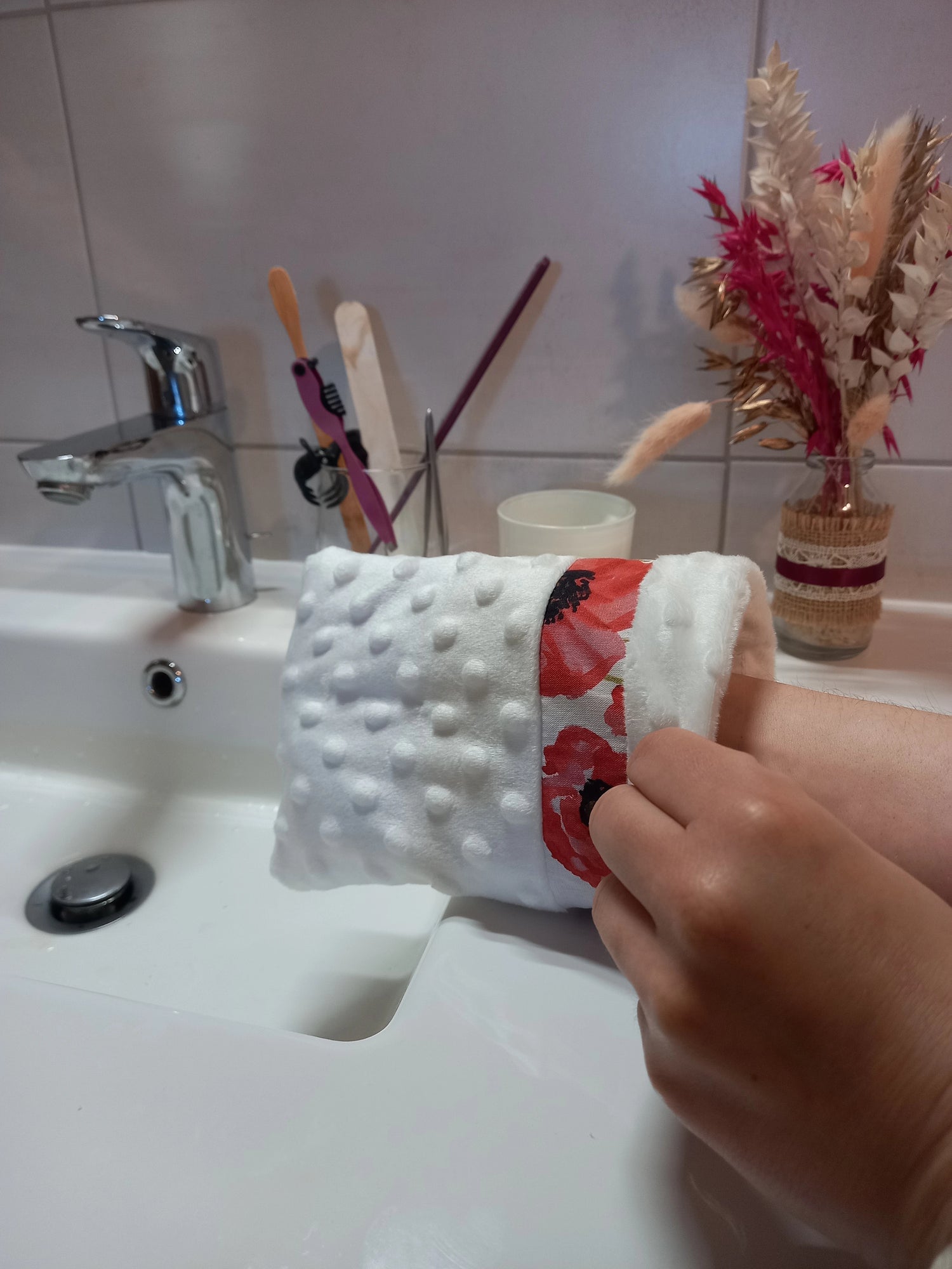 Main portant un gant démaquillant lavable en minky blanc à pois en relief avec sa bande à coquelicots rouges, vue de profil au-dessus du lavabo.