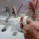 Gant démaquillant lavable en tissu minky blanc à pois en relief, orné d'une bande à coquelicots rouges, enfilé sur une main au-dessus d'un lavabo.