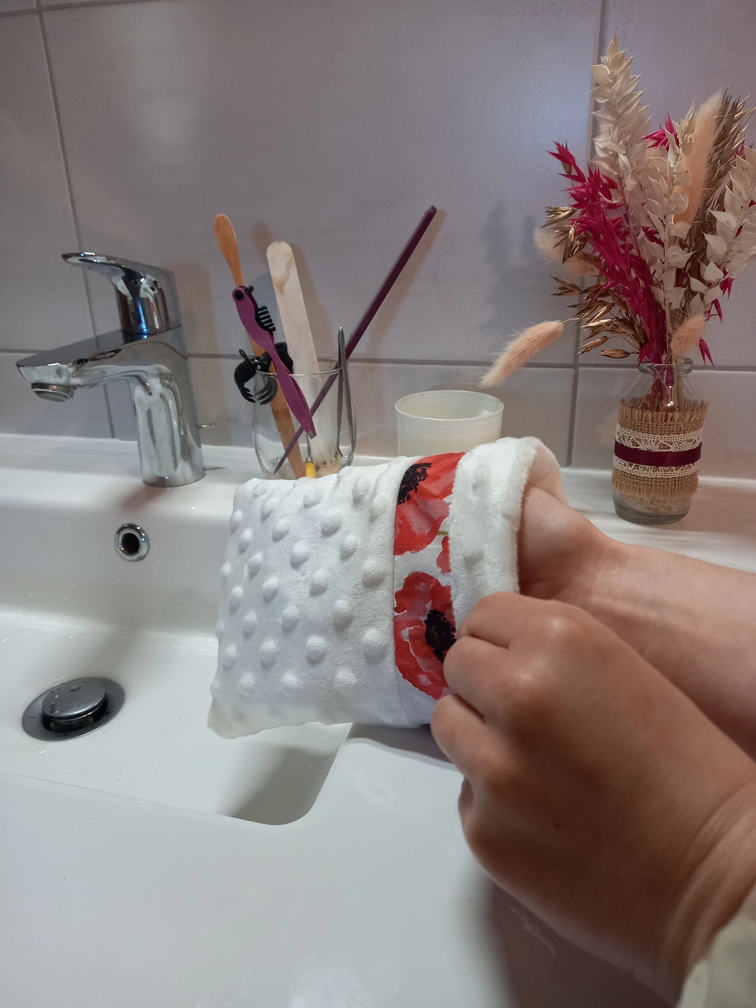 Gant démaquillant lavable en tissu minky blanc à pois en relief, orné d'une bande à coquelicots rouges, enfilé sur une main au-dessus d'un lavabo.