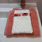 Gant démaquillant lavable en tissu minky blanc à pois en relief, orné d'une bande à coquelicots rouges, posé à plat sur une serviette éponge corail.