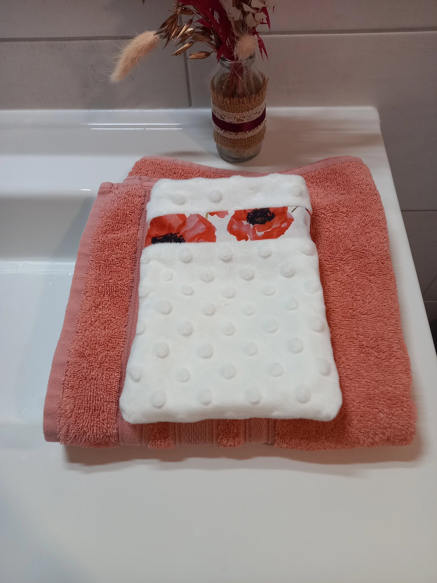 Gant démaquillant lavable en tissu minky blanc à pois en relief, orné d'une bande à coquelicots rouges, posé à plat sur une serviette éponge corail.
