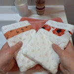 Deux gants démaquillants lavables en minky blanc à pois en relief tenus à deux mains au-dessus d'une serviette éponge corail, l'un orné d'une bande à fleurs printanières orange, l'autre d'une bande à coquelicots rouges.