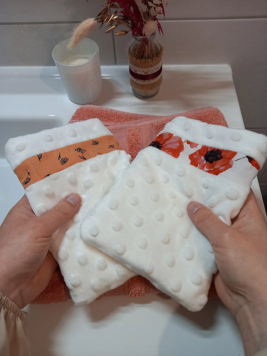 Deux gants démaquillants lavables en minky blanc à pois en relief tenus à deux mains au-dessus d'une serviette éponge corail, l'un orné d'une bande à fleurs printanières orange, l'autre d'une bande à coquelicots rouges.