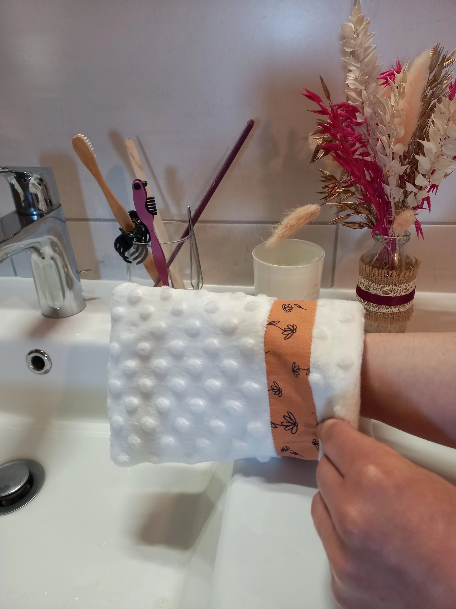 Gant démaquillant lavable en minky blanc à pois en relief à bande fleurs printanières orange, porté sur la main avec le poignet visible, devant un lavabo de salle de bain et un bouquet de fleurs séchées.