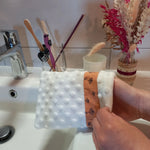 Gant démaquillant lavable en minky blanc à pois en relief, orné d'une bande à fleurs printanières orange, enfilé sur une main au-dessus d'un lavabo de salle de bain, près d'un verre à brosses à dents et d'un bouquet de fleurs séchées.