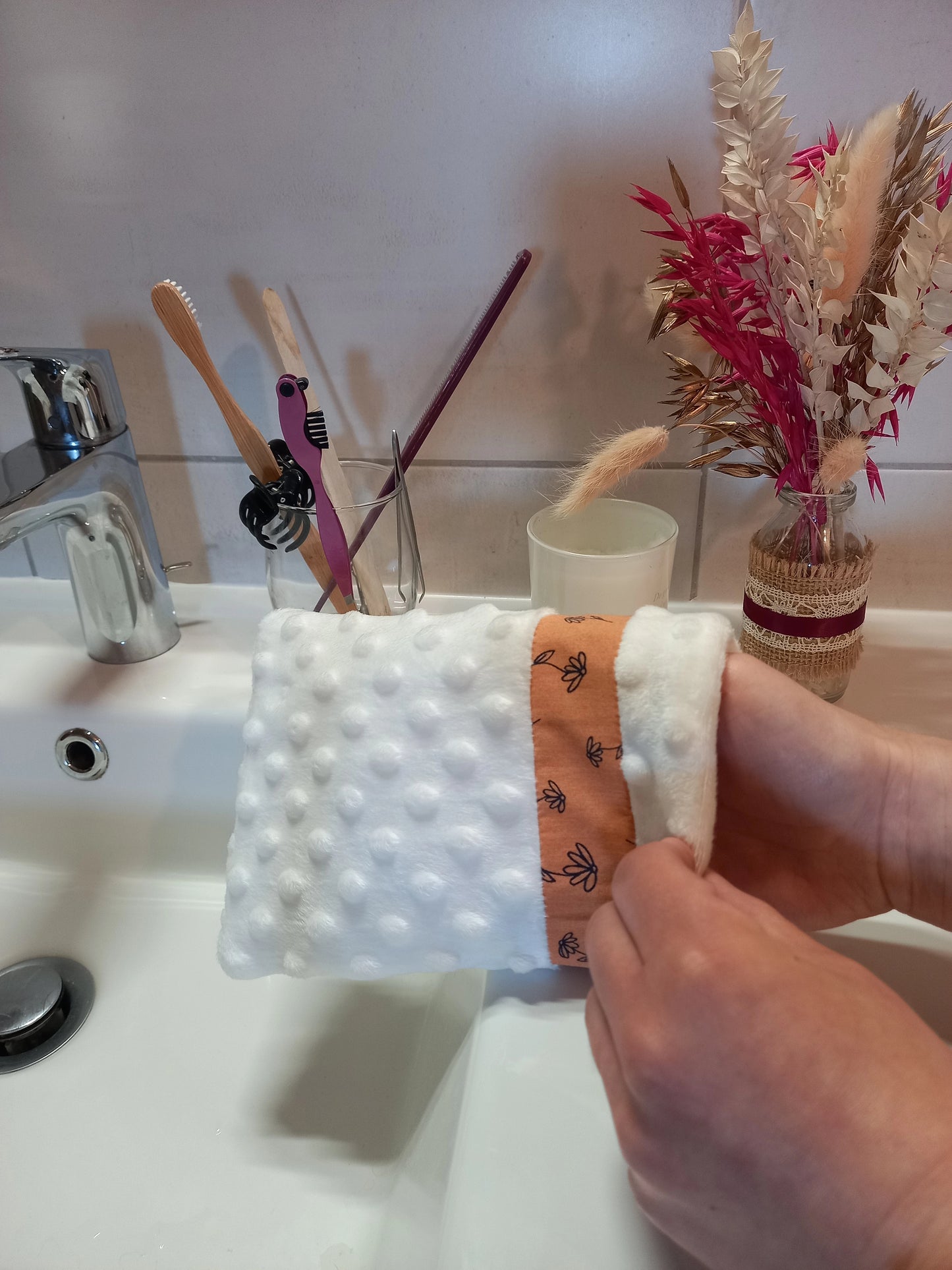 Gant démaquillant lavable en minky blanc à pois en relief, orné d'une bande à fleurs printanières orange, enfilé sur une main au-dessus d'un lavabo de salle de bain, près d'un verre à brosses à dents et d'un bouquet de fleurs séchées.