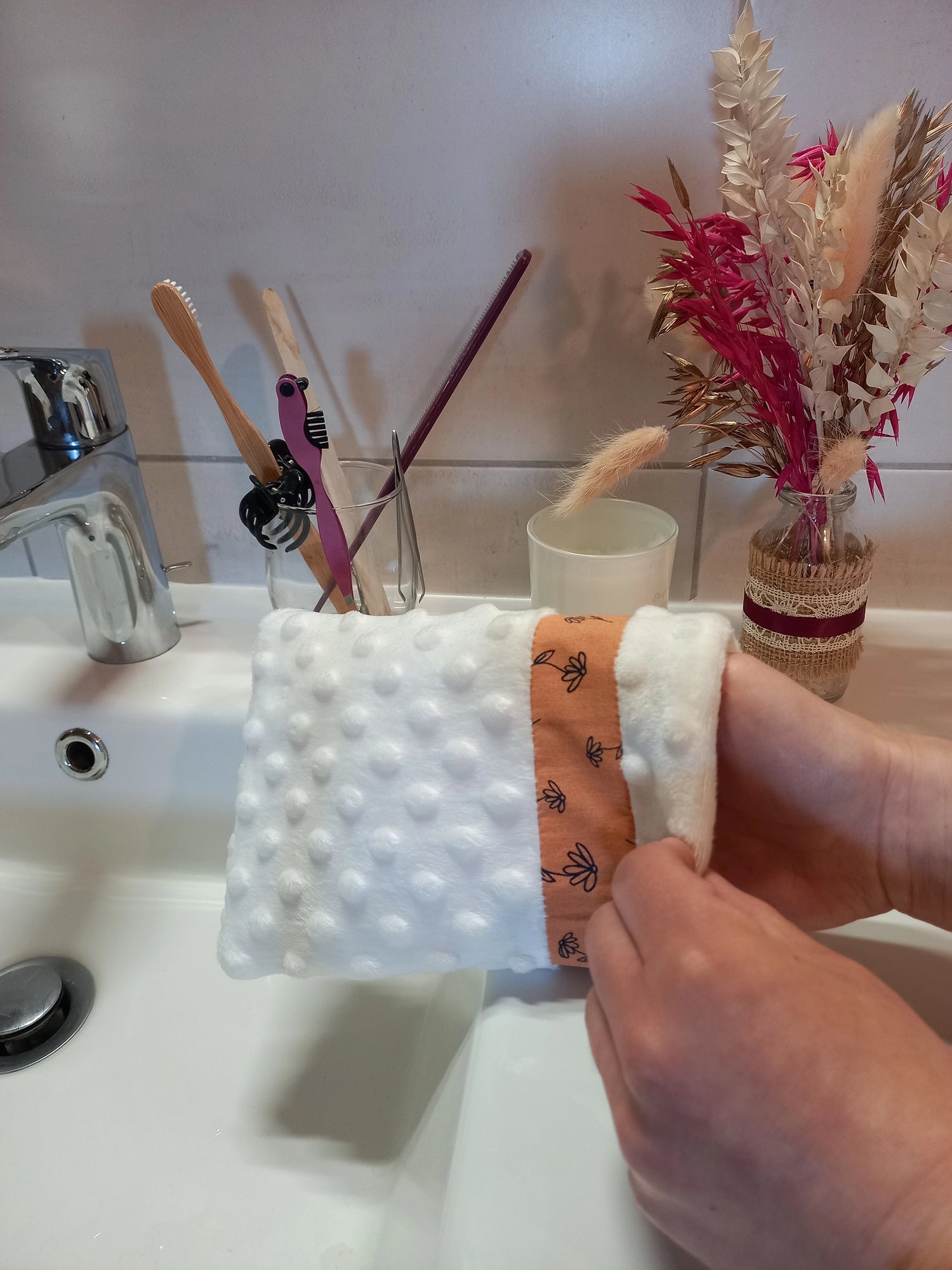 Gant démaquillant lavable en minky blanc à pois en relief, orné d'une bande à fleurs printanières orange, enfilé sur une main au-dessus d'un lavabo de salle de bain, près d'un verre à brosses à dents et d'un bouquet de fleurs séchées.
