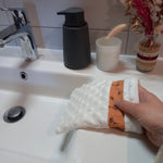 Gant démaquillant lavable en tissu minky blanc à pois en relief, orné d'une bande à fleurs printanières orange, enfilé sur une main au-dessus d'un lavabo.