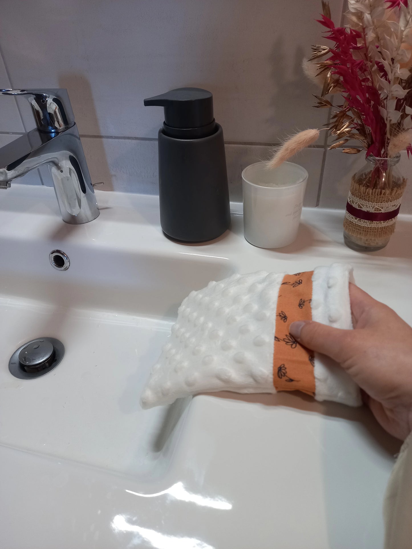 Gant démaquillant lavable en tissu minky blanc à pois en relief, orné d'une bande à fleurs printanières orange, enfilé sur une main au-dessus d'un lavabo.