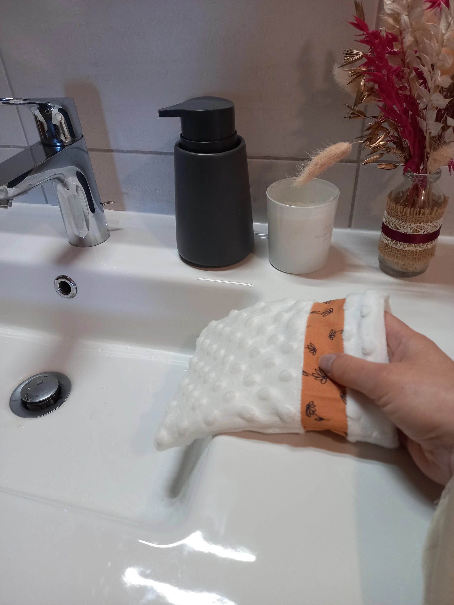 Gant démaquillant lavable en tissu minky blanc à pois en relief, orné d'une bande à fleurs printanières orange, enfilé sur une main au-dessus d'un lavabo.