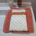 Gant démaquillant lavable en tissu minky blanc à pois en relief, orné d'une bande à fleurs printanières orange, posé à plat sur une serviette éponge corail.