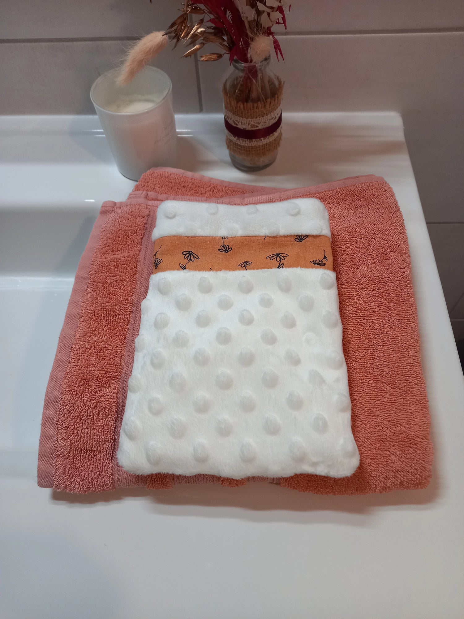 Gant démaquillant lavable en tissu minky blanc à pois en relief, orné d'une bande à fleurs printanières orange, posé à plat sur une serviette éponge corail.