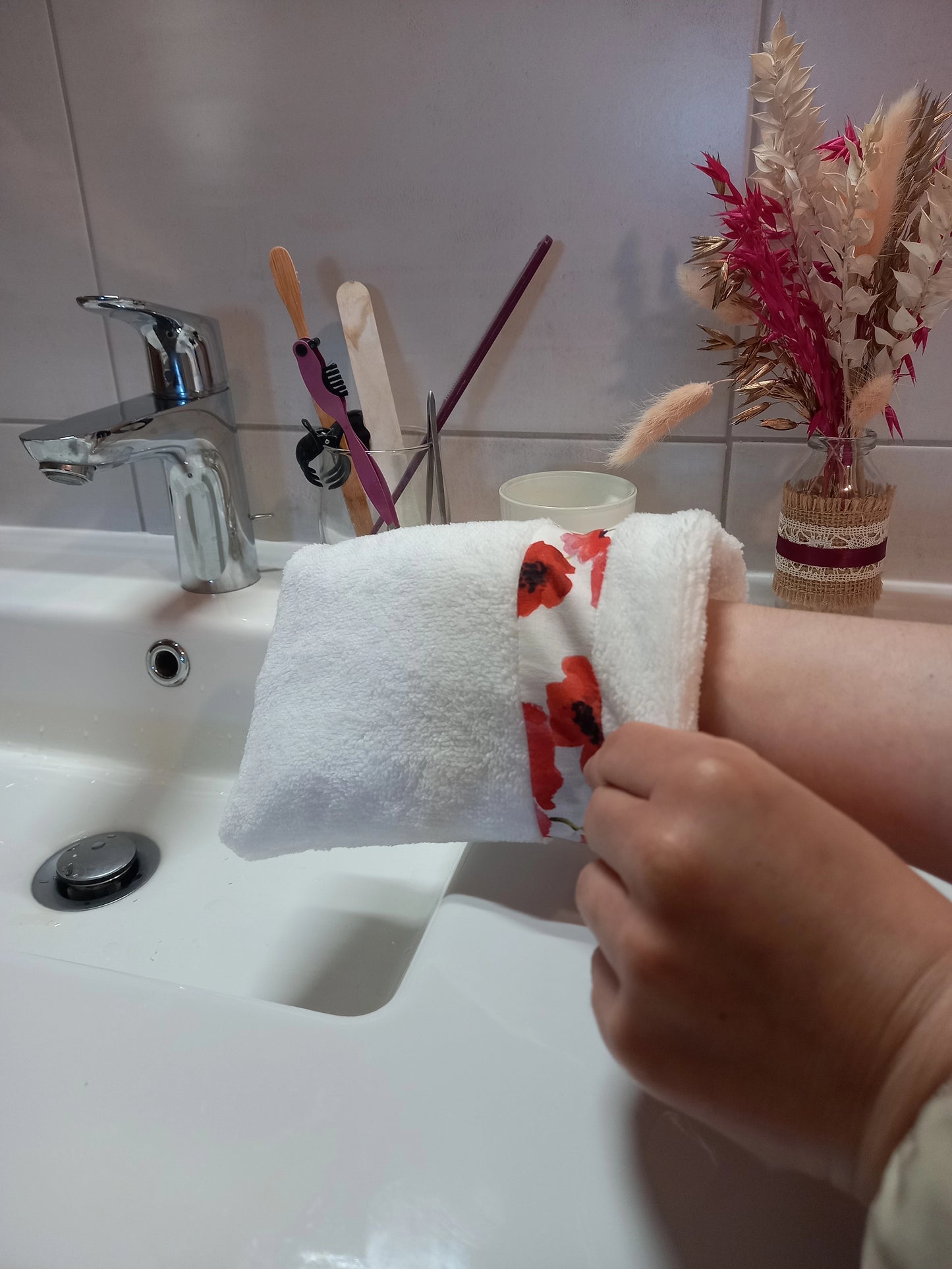 Gant nettoyant visage en éponge blanche à bande de coquelicots rouges porté sur la main avec le poignet visible, au-dessus d'un lavabo de salle de bain, près d'un verre à brosses à dents.