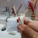 Gant nettoyant visage en éponge blanche à bande de coquelicots rouges enfilé sur la main, vu de près au-dessus d'un lavabo de salle de bain.