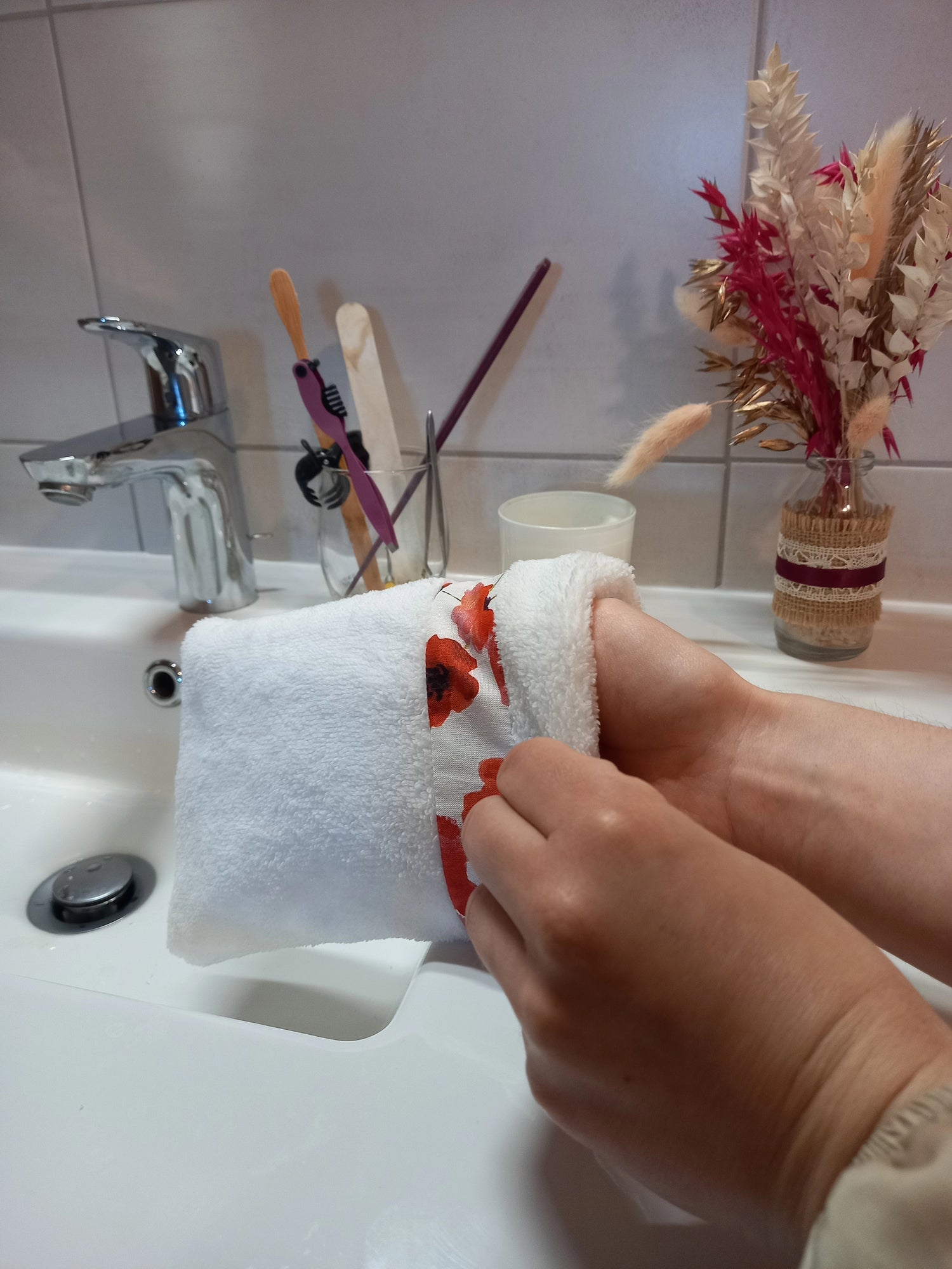 Gant nettoyant visage en éponge blanche à bande de coquelicots rouges enfilé sur la main, vu de près au-dessus d'un lavabo de salle de bain.