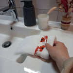 Gant nettoyant visage en éponge blanche à bande de coquelicots rouges enfilé sur une main au-dessus d'un lavabo, près d'un distributeur de savon et d'une bougie.