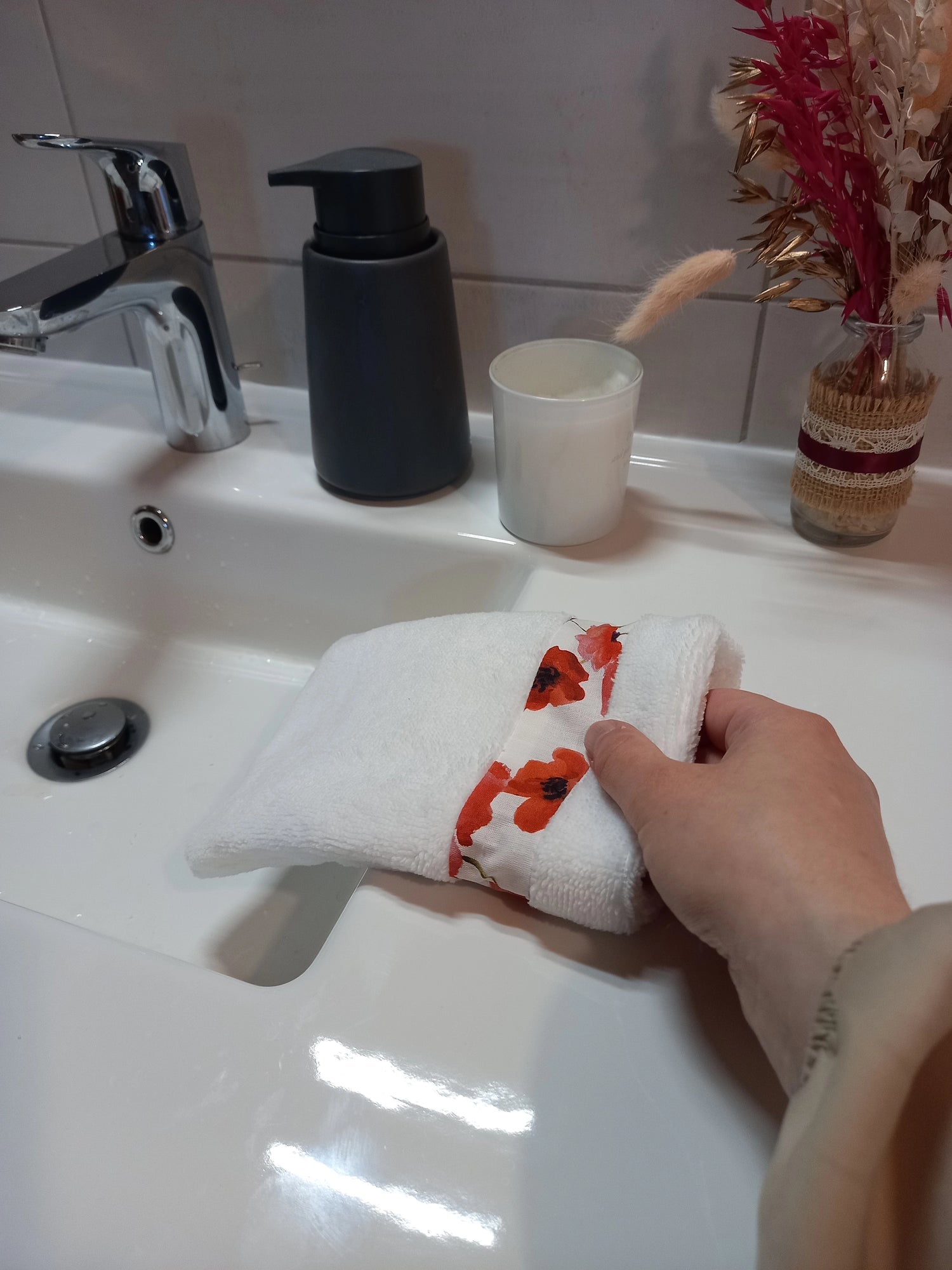 Gant nettoyant visage en éponge blanche à bande de coquelicots rouges enfilé sur une main au-dessus d'un lavabo, près d'un distributeur de savon et d'une bougie.