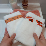 Deux gants nettoyant visage en éponge blanche tenus à deux mains au-dessus d'une serviette éponge corail, l'un orné d'une bande à fleurs printanières orange, l'autre d'une bande à coquelicots rouges.
