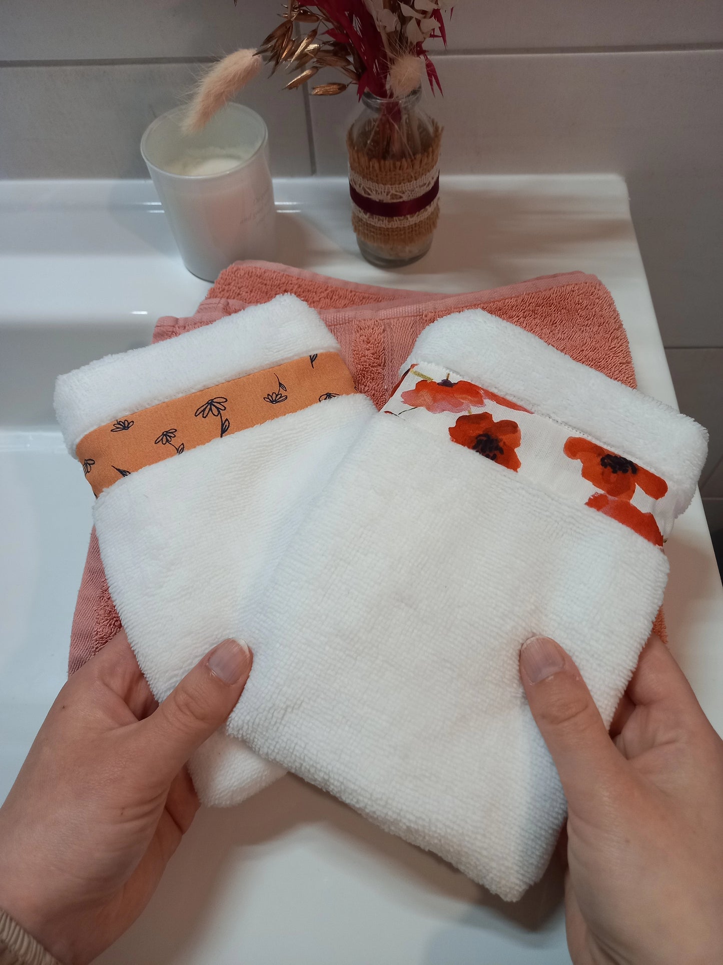 Deux gants nettoyant visage en éponge blanche tenus à deux mains au-dessus d'une serviette éponge corail, l'un orné d'une bande à fleurs printanières orange, l'autre d'une bande à coquelicots rouges.