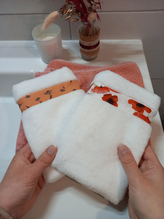Deux gants nettoyant visage en éponge blanche tenus à deux mains au-dessus d'une serviette éponge corail, l'un orné d'une bande à fleurs printanières orange, l'autre d'une bande à coquelicots rouges.
