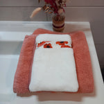 Gant nettoyant visage en éponge blanche, orné d'une bande à coquelicots rouges, posé plié sur une serviette éponge corail, devant un bouquet de fleurs séchées dans une salle de bain.