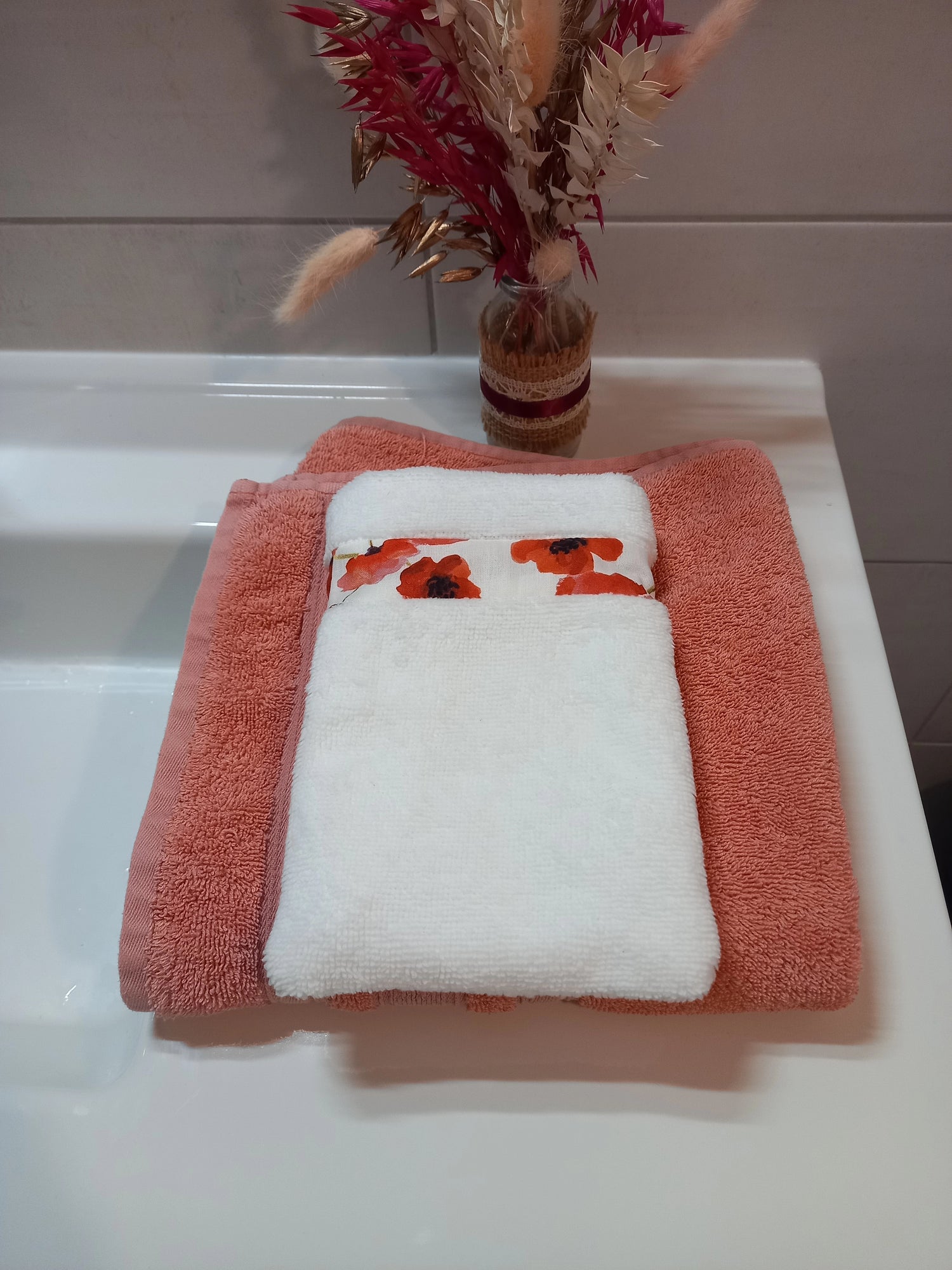 Gant nettoyant visage en éponge blanche, orné d'une bande à coquelicots rouges, posé plié sur une serviette éponge corail, devant un bouquet de fleurs séchées dans une salle de bain.
