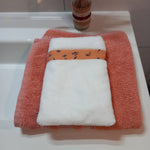 Le gant nettoyant visage de la collection fleurs printanières est posé à plat sur une serviette éponge corail. Il est en tissu éponge blanc avec une bande à motif floral orange.