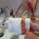 Le gant nettoyant visage fleurs printanières est ajusté à deux mains au-dessus du lavabo. Le tissu éponge blanc et la bande fleurie orange sont visibles.