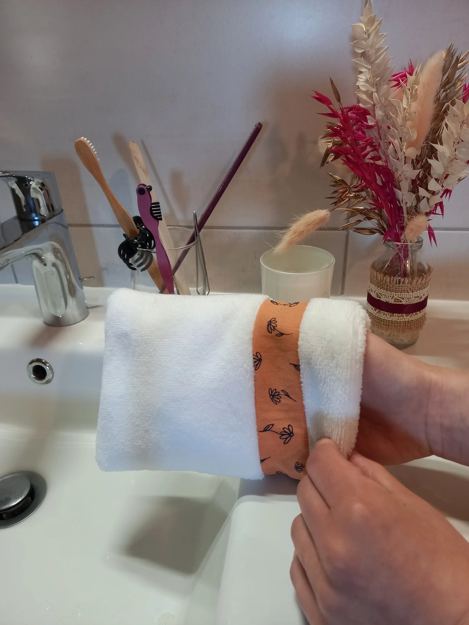 Le gant nettoyant visage fleurs printanières est ajusté à deux mains au-dessus du lavabo. Le tissu éponge blanc et la bande fleurie orange sont visibles.