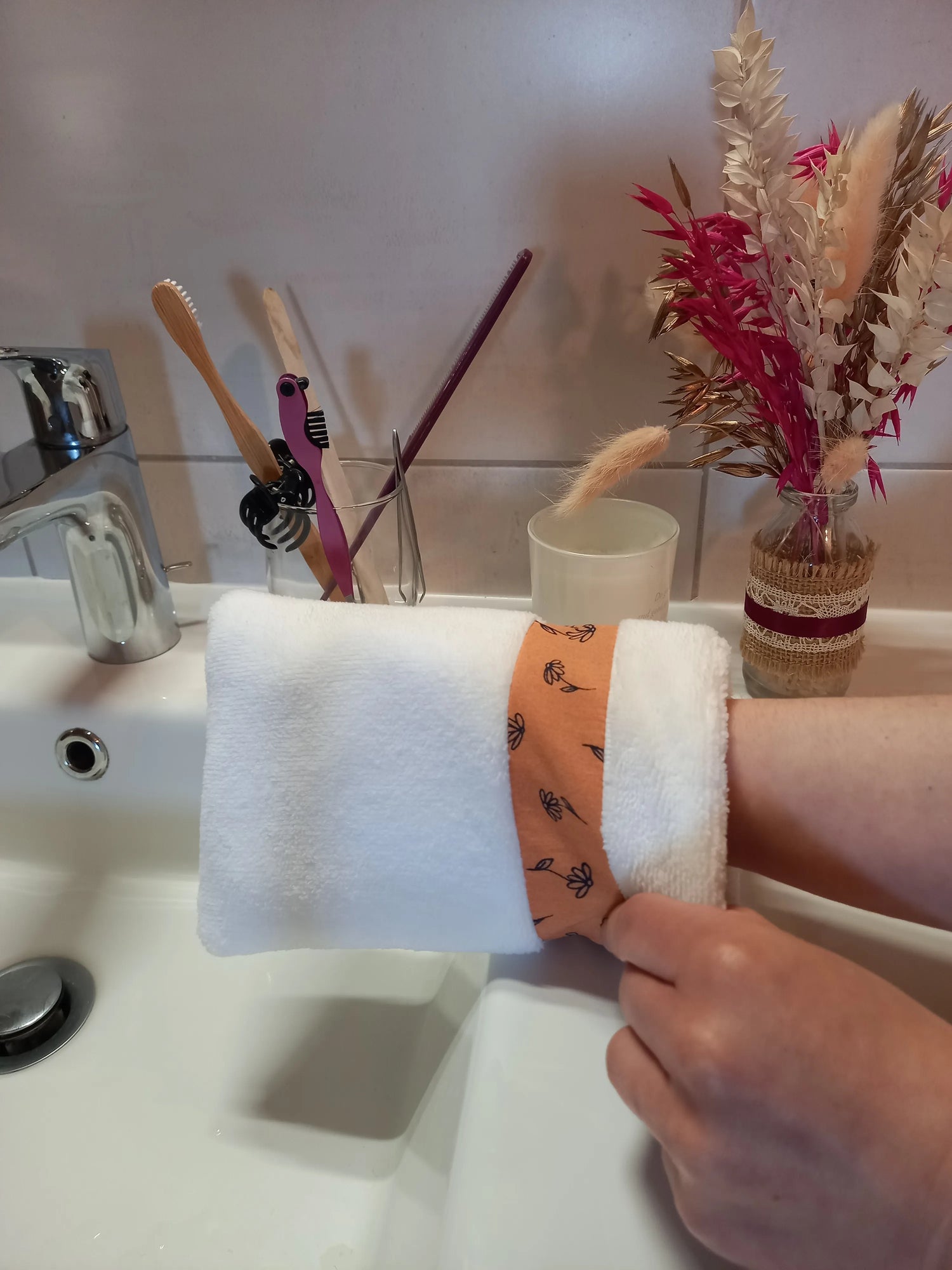 Le gant nettoyant visage fleurs printanières est porté sur une main, tenu près d'un lavabo. La bande à motif floral orange ressort sur l'éponge blanche.