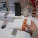 Le gant nettoyant visage fleurs printanières est enfilé sur une main et posé sur le rebord d'un lavabo. Le tissu éponge blanc et la bande à fleurs orange sont visibles.