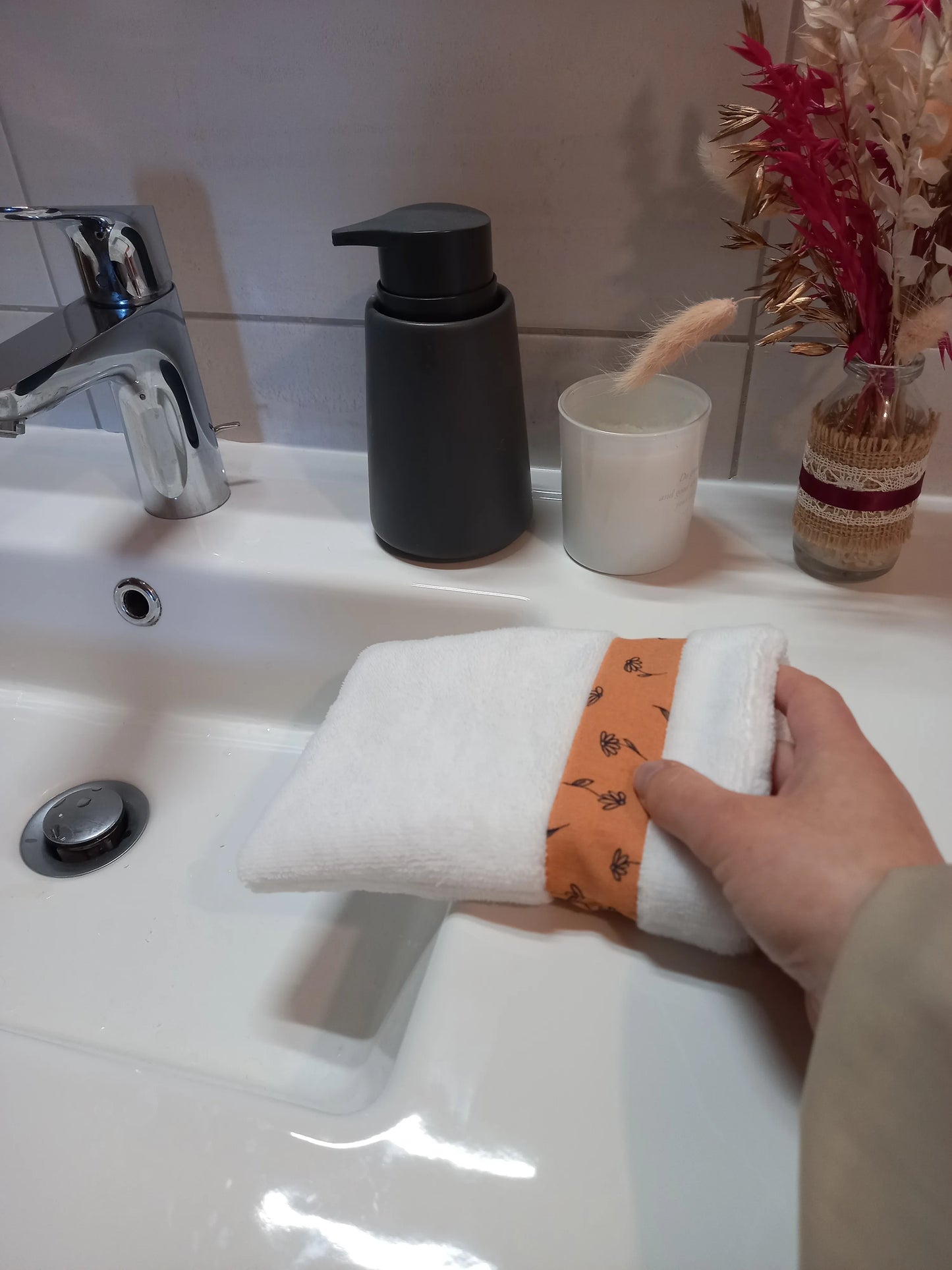Le gant nettoyant visage fleurs printanières est enfilé sur une main et posé sur le rebord d'un lavabo. Le tissu éponge blanc et la bande à fleurs orange sont visibles.