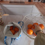 Pochon alimentaire motif fleuri bleu rempli d'oranges posé près d'une corbeille à fruits et une main saisissant une clémentine