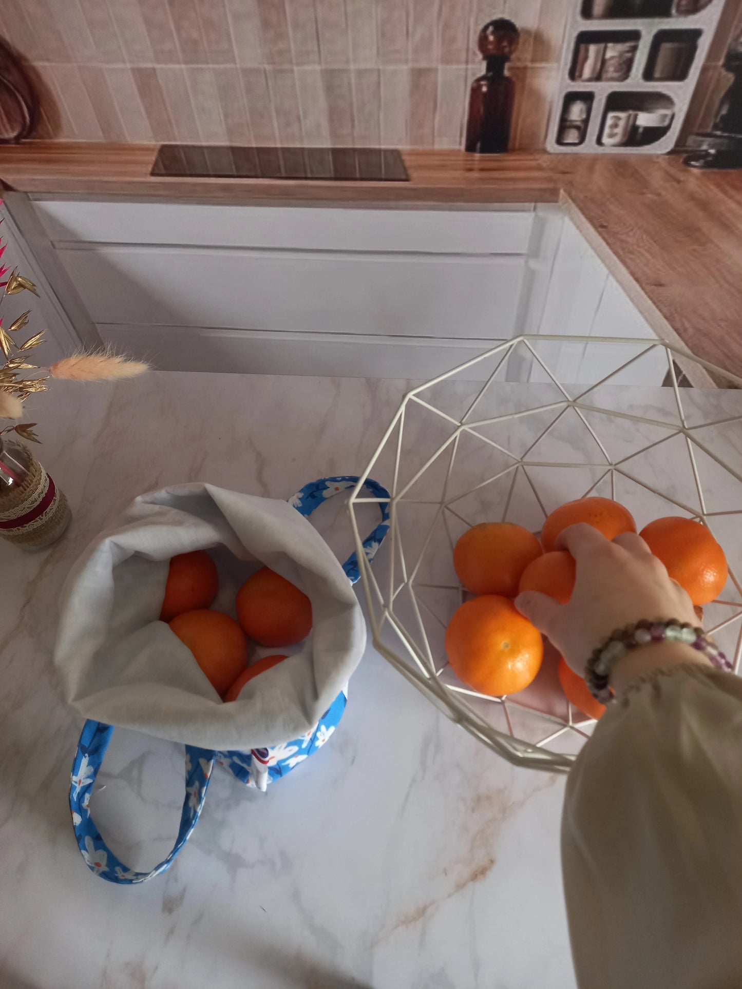 Pochon alimentaire motif fleuri bleu rempli d'oranges posé près d'une corbeille à fruits et une main saisissant une clémentine