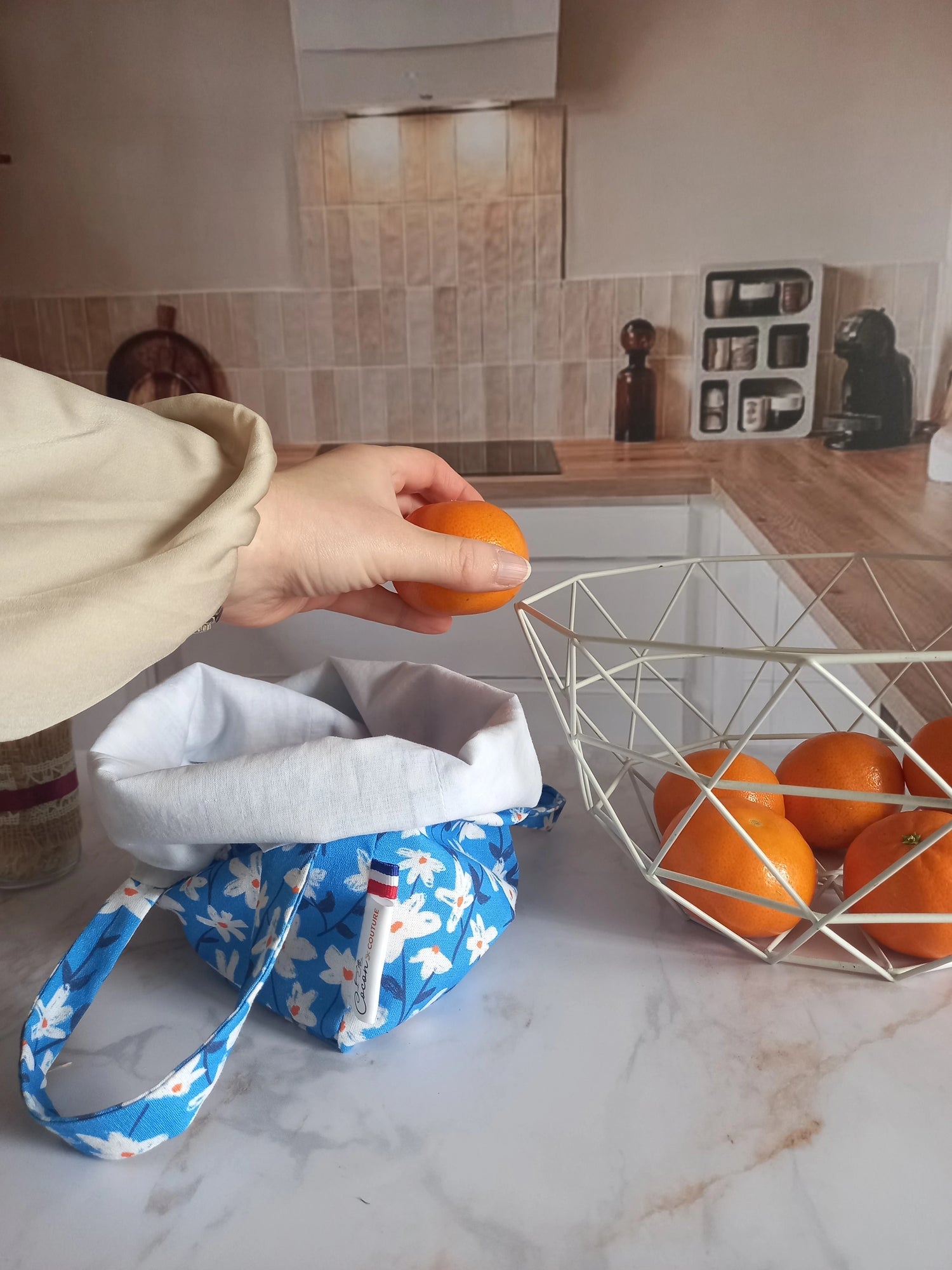 Pochon alimentaire en coton motif fleuri bleu avec l'étiquette Cocon Couture et une main y déposant une orange