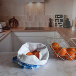 Pochon alimentaire en coton motif fleuri bleu rempli d'oranges, posé sur un plan de travail en marbre à côté d'une corbeille à fruits
