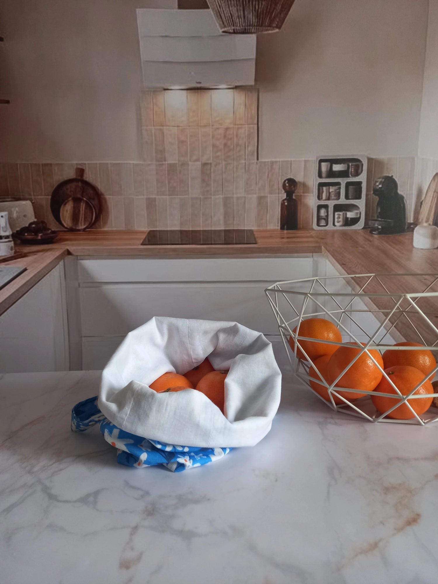 Pochon alimentaire en coton motif fleuri bleu rempli d'oranges, posé sur un plan de travail en marbre à côté d'une corbeille à fruits