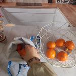 Main déposant une orange dans un pochon alimentaire motif fleuri bleu rempli de fruits, vue de dessus