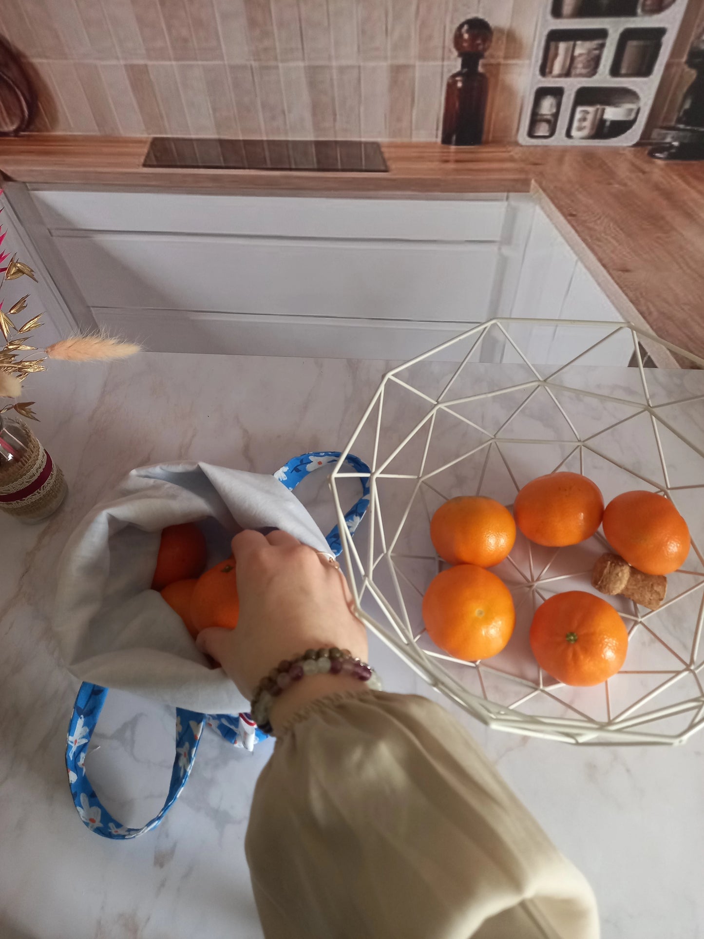 Main déposant une orange dans un pochon alimentaire motif fleuri bleu rempli de fruits, vue de dessus