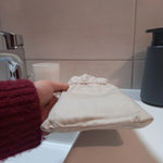 Pochon en lin beige des carrés démaquillants lavables, fermé par son cordon coulissant et tenu à la main sur le rebord d'un lavabo.