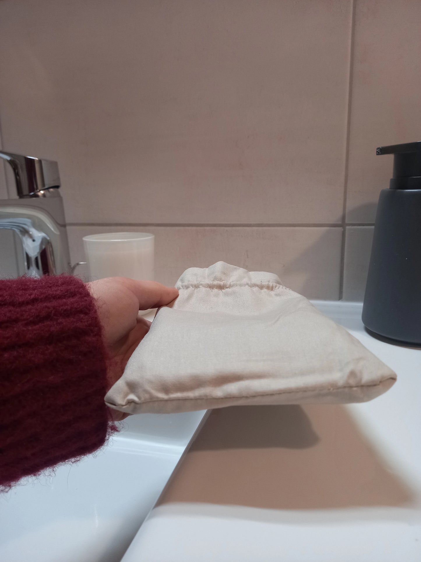 Pochon en lin beige des carrés démaquillants lavables, fermé par son cordon coulissant et tenu à la main sur le rebord d'un lavabo.