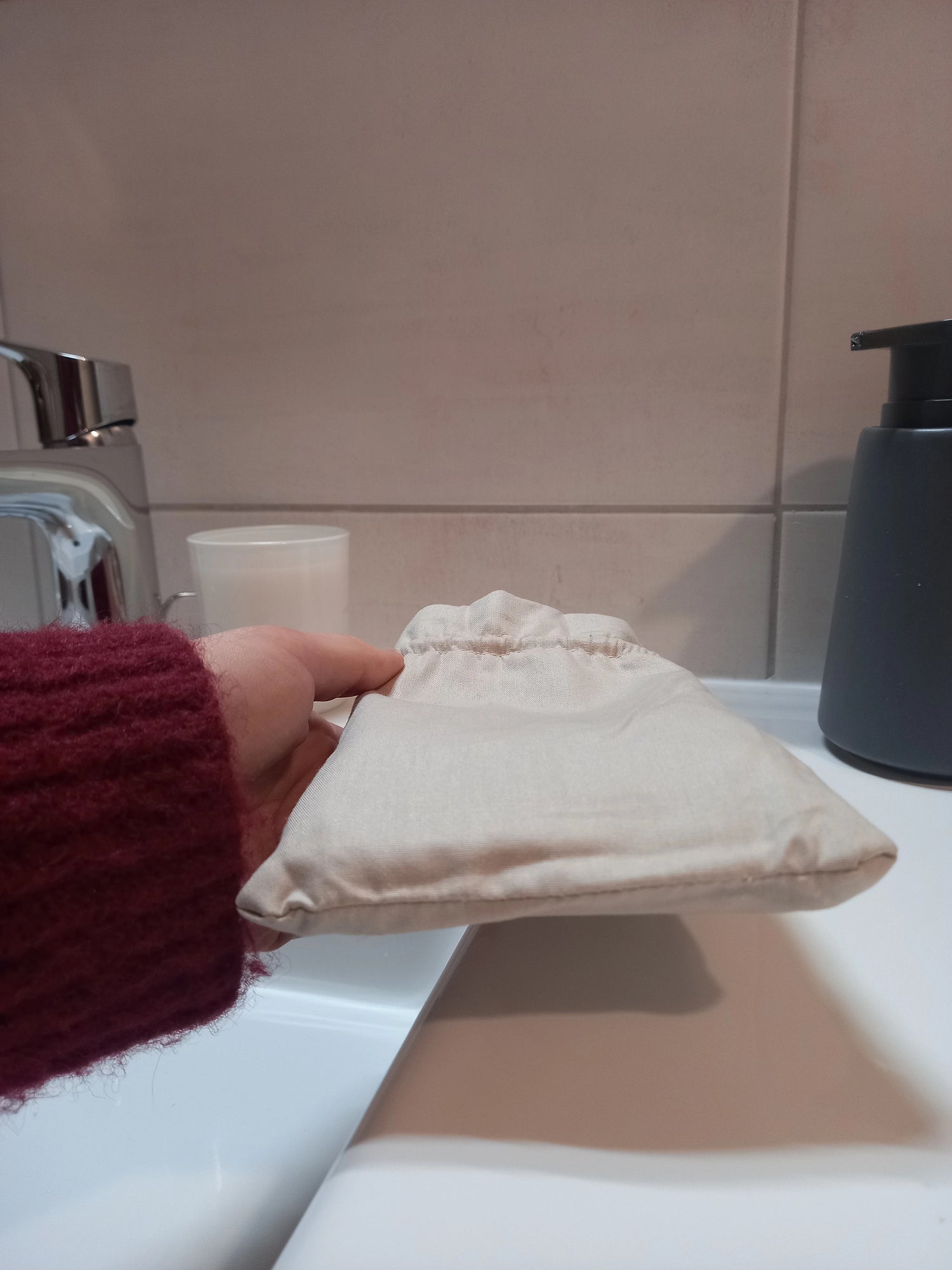 Pochon en lin beige des carrés démaquillants lavables, fermé par son cordon coulissant et tenu à la main sur le rebord d'un lavabo.