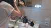 Devant son miroir, une femme se démaquille le visage avec un carré démaquillant lavable, le pochon et les carrés Cocon Couture sont posés sur le lavabo.