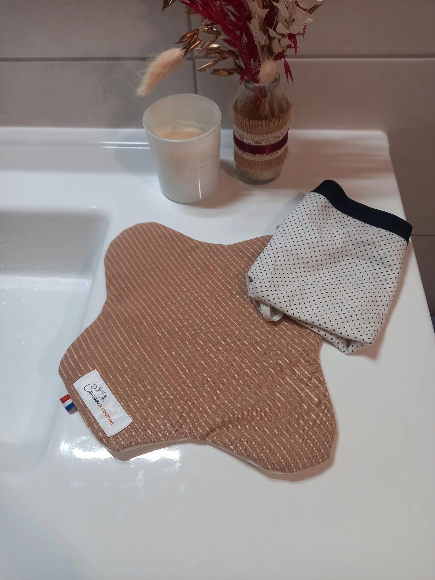 Le protège-slip en coton lavable à rayures beiges est plié et fermé par son bouton-pression, tenu dans une main, l'étiquette Cocon Couture étant visible.