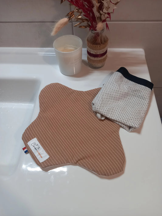 Le protège-slip en coton lavable à rayures beiges est plié et fermé par son bouton-pression, tenu dans une main, l'étiquette Cocon Couture étant visible.