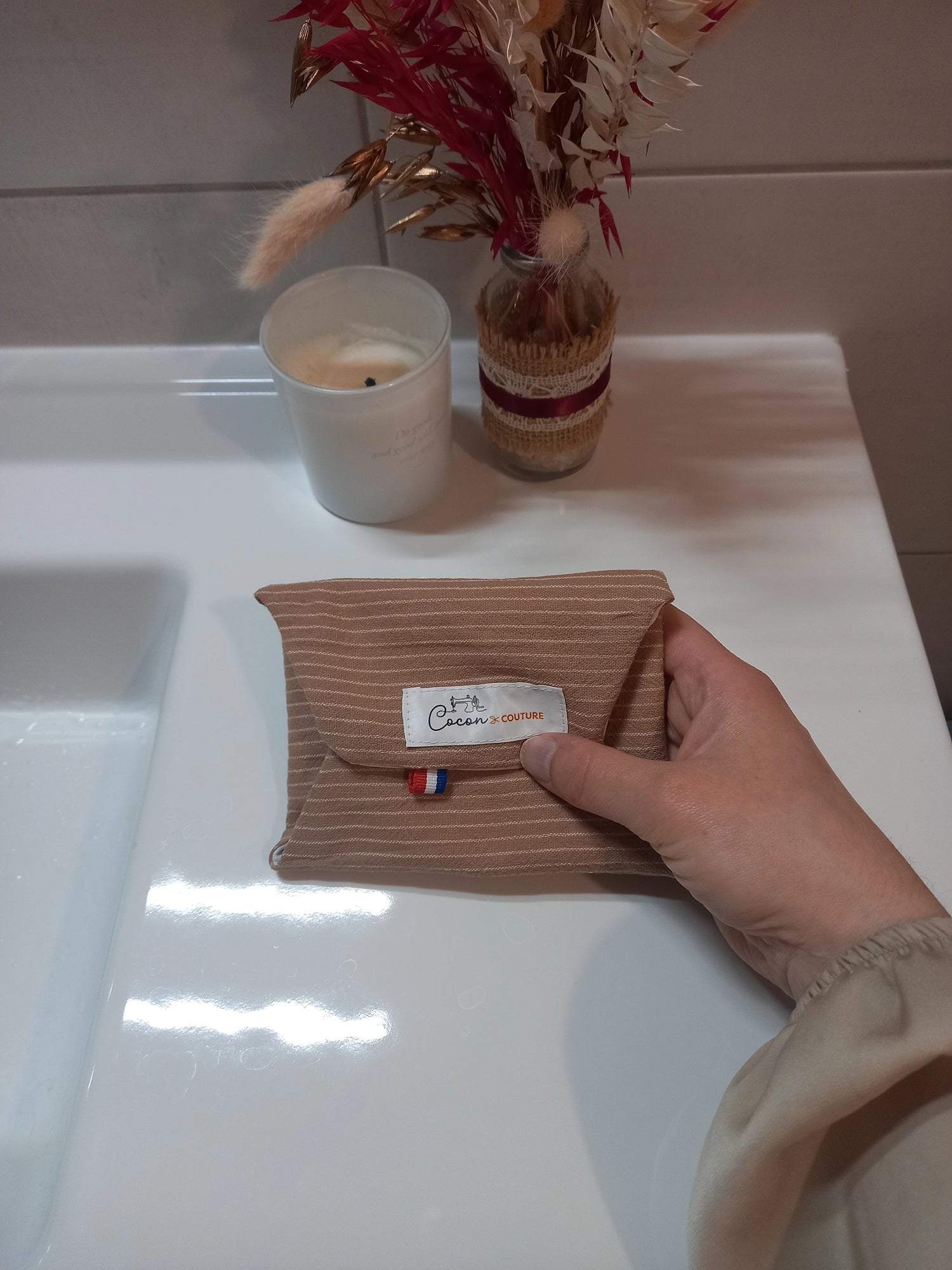 Le protège-slip en coton lavable à rayures beiges est plié et fermé par son bouton-pression, tenu dans une main, l'étiquette Cocon Couture étant visible.