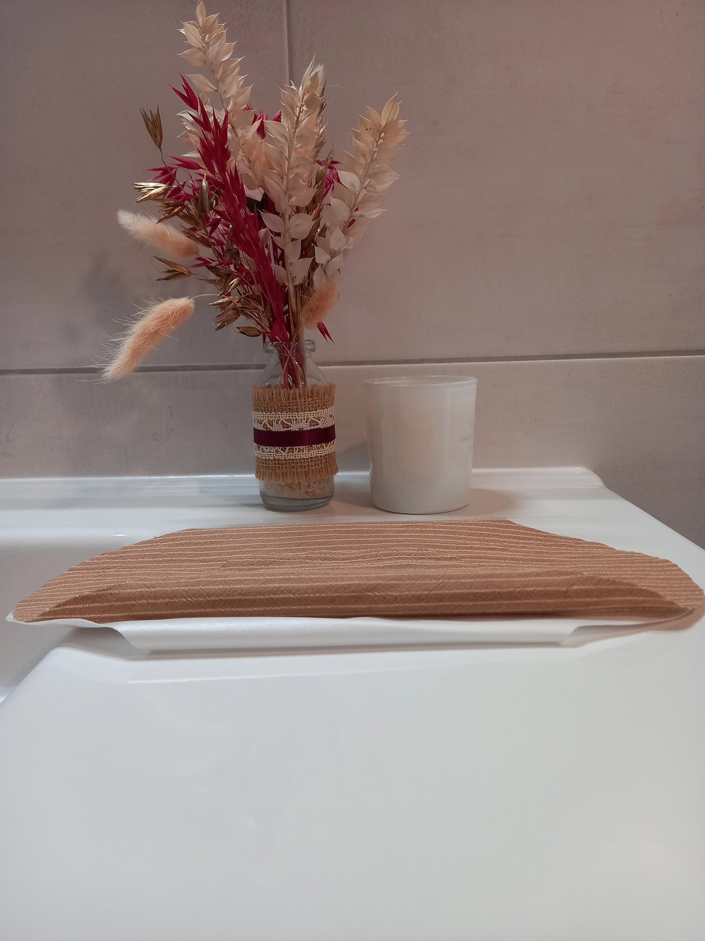 Le protège-slip en coton lavable à rayures beiges est posé plié sur le rebord d'un lavabo, près d'une bougie et d'un bouquet de fleurs séchées.