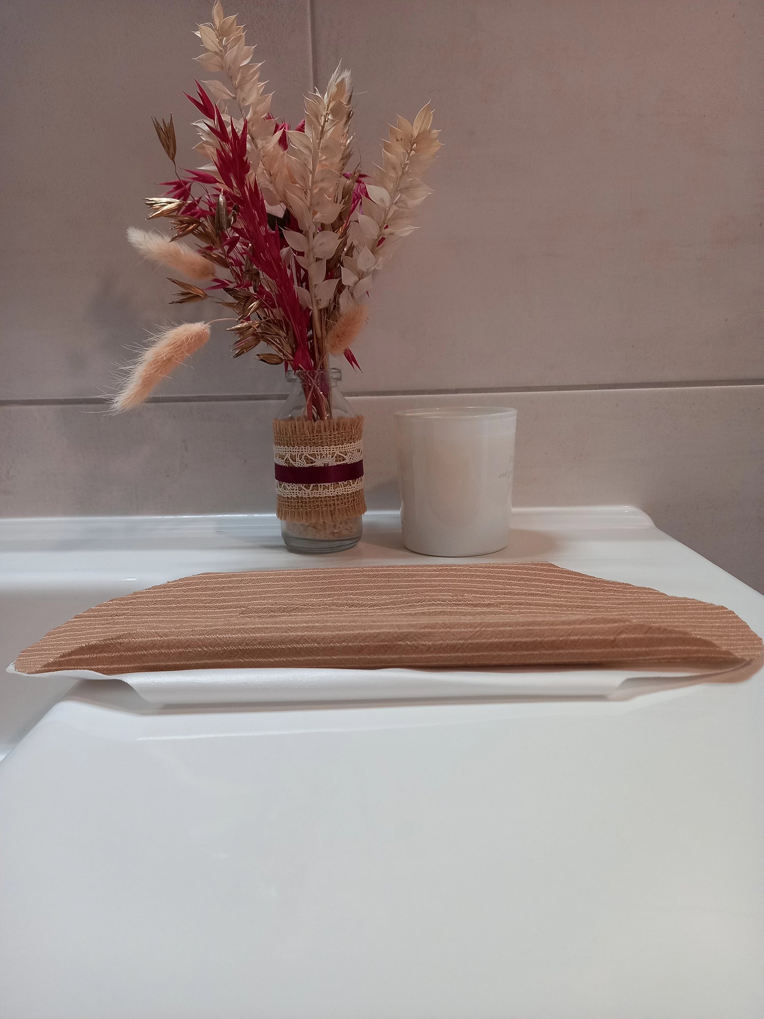 Le protège-slip en coton lavable à rayures beiges est posé plié sur le rebord d'un lavabo, près d'une bougie et d'un bouquet de fleurs séchées.