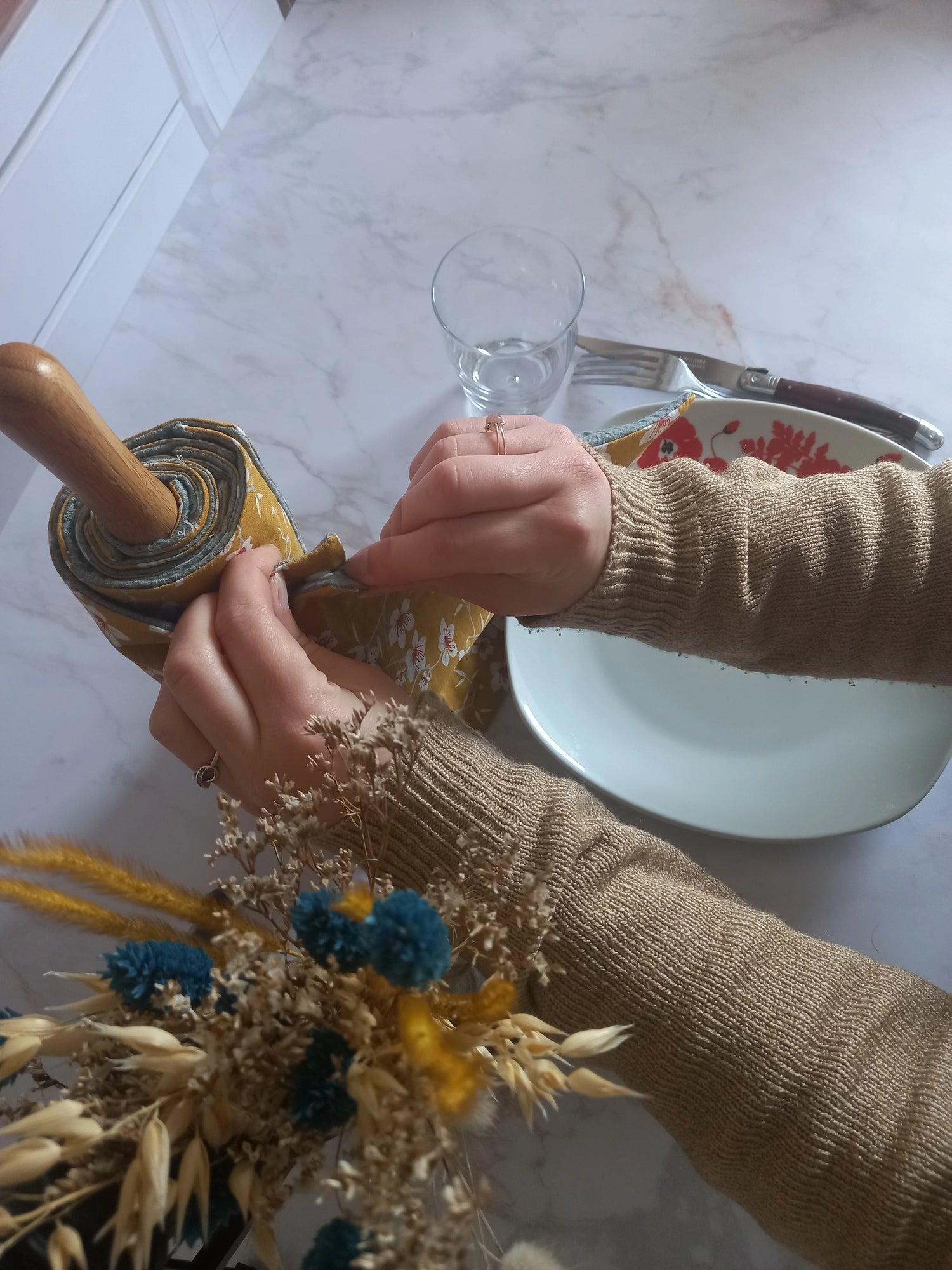 Deux mains déroulant un rouleau essuie-tout lavable Cerisier sur un plan de travail avec, à côté, un bouquet de fleurs séchées